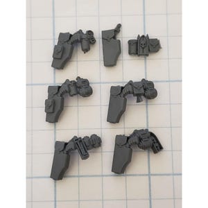 May include: A collection of small, gray plastic model components. The pieces include what appear to be gun-like structures, pouches, and shield-like elements. The parts are arranged on a grid-patterned surface, suggesting they are for a miniature model or game.