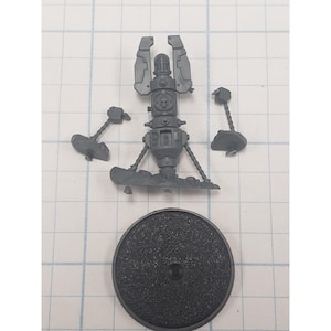 May include: A gray plastic miniature model kit, featuring a central structure with a skull emblem, two hammer-like components, and a textured circular base. The model is designed for assembly.