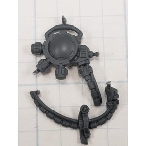 May include: A dark gray plastic miniature weapon component, featuring a circular central body with protruding details, including knobs and a handle-like structure. A separate curved piece with a skull-like emblem is also present.