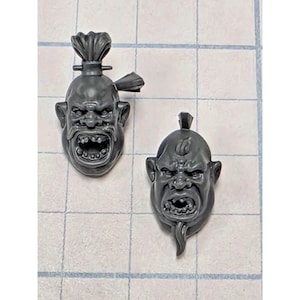 May include: Two unpainted grey plastic miniature heads featuring orc-like facial features with wide, open mouths showing teeth. The left head has a topknot hairstyle, while the right head features a small mohawk and a pointed chin beard.