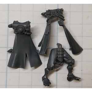 May include: Gray plastic model kit pieces for a sci-fi character. The pieces include a cape, legs, and a belt with a detailed design.