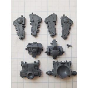 May include: A collection of gray plastic model kit parts, including various armor pieces and mechanical components. The parts are arranged on a grid-lined surface, showcasing different shapes and sizes, ideal for miniature wargaming or model building.