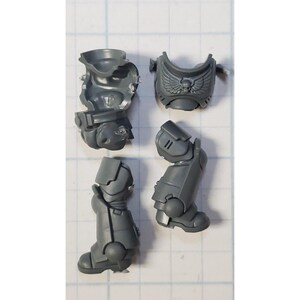May include: A collection of unpainted, gray plastic miniature parts for a tabletop game. The components include a torso, chest piece with a winged emblem, and two leg sections. The pieces are designed to be assembled.