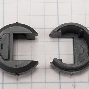 Puede incluir: Componente de pl&aacute;stico gris oscuro de dos piezas. Cada pieza tiene una forma exterior semicircular con una secci&oacute;n rectangular. Una pieza tiene una peque&ntilde;a incrustaci&oacute;n rectangular. Las piezas est&aacute;n separadas.