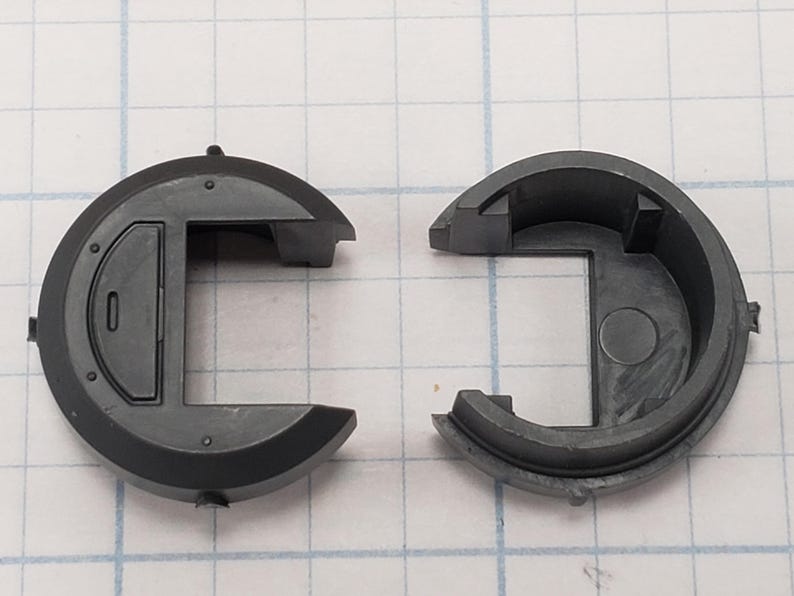 Puede incluir: Componente de pl&aacute;stico gris oscuro de dos piezas. Cada pieza tiene una forma exterior semicircular con una secci&oacute;n rectangular. Una pieza tiene una peque&ntilde;a incrustaci&oacute;n rectangular. Las piezas est&aacute;n separadas.