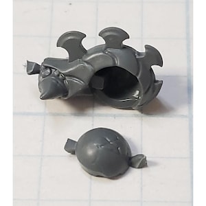 May include: A gray plastic miniature model component, possibly for a tabletop game. The piece has a rounded, helmet-like structure with protruding spikes and fins. A separate, smaller, rounded piece is also present.