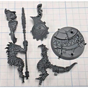 May include: A set of unpainted gray plastic miniature model parts including a reptilian creature torso with a spear, a separate lower body section, a circular shield with geometric carvings, and a round textured base with cracked stone details.