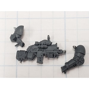 May include: A gray plastic miniature model of a weapon with attached scope and two arm pieces. The weapon has intricate details and is designed for tabletop gaming or model building.