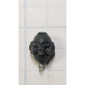 May include: A gray plastic miniature helmet with a skull face design. The helmet has a rounded top, two side attachments, and a small base. The skull features eye sockets, a nose cavity, and teeth. The miniature is likely for a tabletop game.