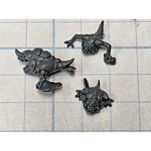 May include: Three unpainted dark gray plastic miniature creature components. One larger figure features a horned, armored back and tail. A second figure is depicted in a crawling pose with limbs extended. A third smaller piece shows a detailed head.