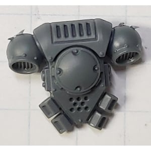 May include: A gray plastic miniature of a mechanical backpack with two spherical side components and a central, rounded section. The backpack has a series of rectangular vents and a circular, riveted plate. The bottom features a triangular design with multiple rectangular protrusions.