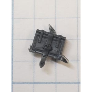May include: A small, dark gray miniature chest with a hinged lid and a clasp. The chest has two decorative spikes on the sides and is likely a component of a model or miniature figure.