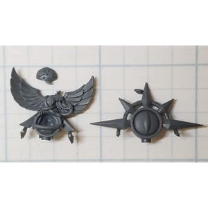 May include: Two gray plastic miniature figurines for a tabletop game. One figurine is a winged creature holding a sphere. The other figurine is a star-shaped object with a central eye.