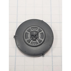 May include: A gray, circular plastic component featuring a skull and crossbones emblem at its center. The emblem is surrounded by a circular design with three small circles at the top and bottom. The component has a series of small rivets around its edge.