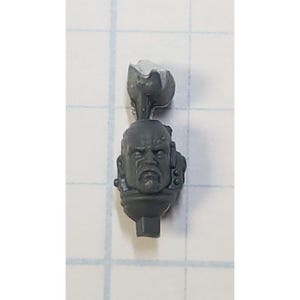 May include: A gray plastic miniature head with a helmet and a stern expression. The head is designed for use in a tabletop wargame.
