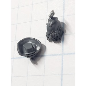 May include: Two gray plastic miniature components. One is a hat-like piece with a square base, and the other is a detailed head with a stylized face and a pointed top. The pieces are on a grid background.
