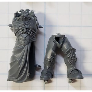 May include: Gray plastic miniature model of a space marine with a detailed torso and legs. The torso features a sculpted eagle and a cape. The legs have sculpted armor and boots.