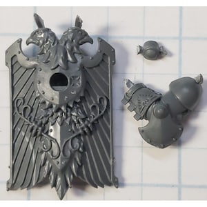 May include: A gray plastic miniature wargaming model of a shield with a double-headed eagle design. The shield has a central circular opening and a decorative border. The eagle has a detailed design with feathers and a crest.