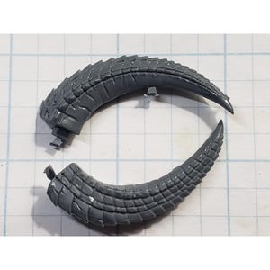 May include: Two gray, curved, segmented plastic horns, possibly for a figurine or model. The horns have a textured, scale-like pattern and are positioned on a grid background. One horn is broken.