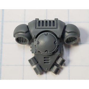 May include: A gray plastic miniature of a space marine's power armor back and shoulder assembly. The piece features a central circular component, two shoulder pads with vents, and a lower section with three rectangular details. The overall design is detailed and mechanical.