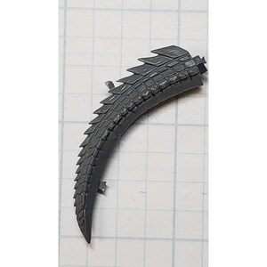 May include: A curved, gray plastic tail segment with a textured, scaled pattern. The tail has a series of pointed dorsal fins along its top edge. The object is likely a component of a model or toy.