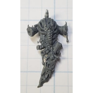 May include: A gray plastic miniature figure, possibly for tabletop gaming. The figure has a detailed, mechanical design with tubes, armor plating, and a large, ornate blade. The figure is posed in a dynamic stance, suggesting action.