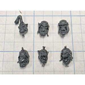 May include: Six unpainted gray plastic miniature orc heads arranged in two rows. The heads feature various expressions, including open mouths with teeth, and are wearing different styles of helmets, including one with a topknot and one with a monocle.