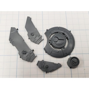 May include: A collection of unassembled, gray plastic model kit parts. The components include angular panels, a circular piece with a central design, and a small cylindrical piece. The parts are arranged on a grid-lined surface.