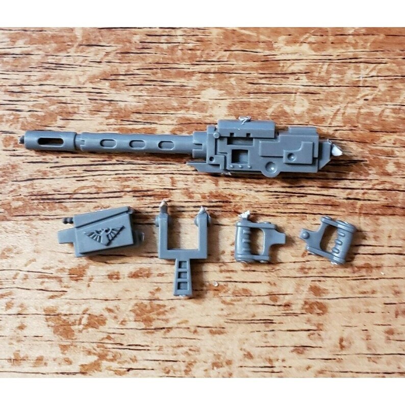 Op de afbeelding: Een verzameling grijze plastic miniatuurwapenonderdelen, waaronder een lange loop met cirkelvormige openingen, een rechthoekige behuizing en verschillende kleinere componenten. De stukken zijn op een licht oppervlak gerangschikt.