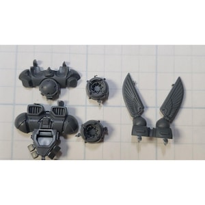 May include: A set of gray plastic miniature parts for a model kit. The parts include a torso, a backpack, two wings, and two round pieces.