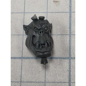 May include: A dark gray plastic miniature head of an orc character featuring prominent tusks, a furrowed brow, and a circular mechanical eyepiece over the left eye. The head is sculpted with pointed ears and a small protrusion at the top and bottom.