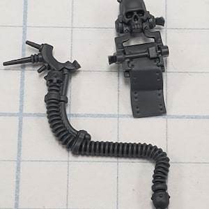 May include: Two dark gray plastic model components. One is a flexible, ribbed tube with skull details and a small skull at the end. The other is a rectangular piece with a skull on top. Both are detailed with mechanical elements.