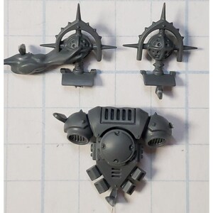 May include: A set of gray plastic miniature parts for a model kit. The parts include a backpack, two arm pieces, and two head pieces.