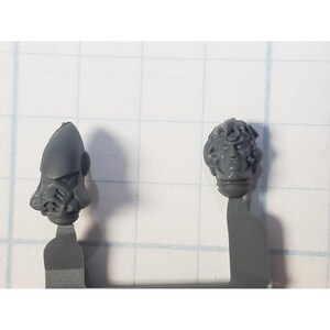 May include: Two plastic miniature heads for a model kit. One head is a helmeted warrior with a face mask. The other head is a bare-headed warrior with a detailed face.