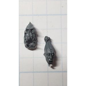 May include: Two gray plastic miniature heads, one with a closed mouth and the other with an open mouth, for use in a tabletop game or hobby project.
