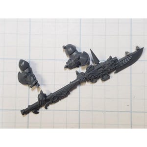 May include: A gray plastic miniature weapon with ornate details, including a spear-like shaft, a blade with wing-like embellishments, and two armor pieces. The components are unassembled and lie on a grid-patterned surface.