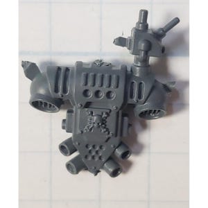 May include: A gray plastic miniature model component, featuring a central rectangular structure with multiple circular and rectangular openings. It has several protruding tubes and a shield-shaped element with a skull emblem, likely for a tabletop game.