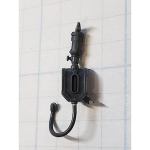 May include: A black plastic model of a sci-fi weapon with a hose and a small tank. The tank has a rectangular shape with a round opening in the center.