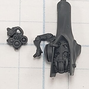 May include: Two-piece, gray plastic miniature model kit components. One piece features a tall, hooded structure with a detailed face and mechanical elements. The other piece is a smaller, multi-part component with circular details and a mechanical arm.