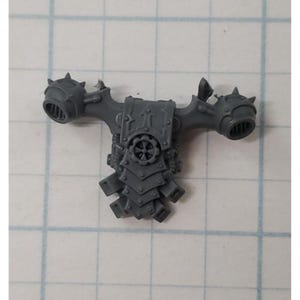 May include: A gray plastic miniature figure component. The piece features two spiked, cylindrical protrusions on either side of a central structure with a gear-like emblem. The design includes layered, angular armor-like details.