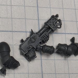 May include: A dark gray plastic miniature weapon component featuring a heavy firearm with a central emblem, accompanied by two separate arm segments with shoulder pauldrons, all cast in unpainted molded plastic for assembly in tabletop gaming.