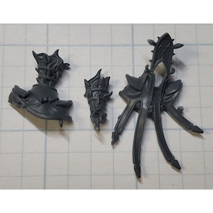 May include: Three gray plastic pieces of a miniature model kit. The pieces are detailed with sharp edges and spikes. The pieces are for a fantasy or science fiction model.