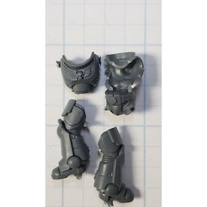 May include: A collection of gray plastic miniature model parts, including a chest piece with a skull and wing emblem, a torso, and two leg pieces. The parts are designed for assembly and are likely for a tabletop game.