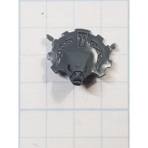 May include: A gray plastic miniature component featuring a boar's head within a cogwheel design. The boar's face has detailed features, and the cogwheel has intricate patterns. This piece is likely for a tabletop game or model.
