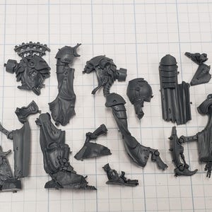 May include: A collection of unassembled, gray plastic model kit pieces, including armor, limbs, and a head with intricate details. The parts are arranged on a grid-lined surface, suggesting a miniature figure or vehicle.