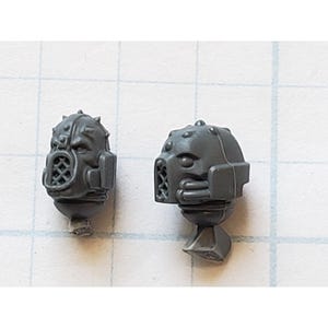 May include: Two gray plastic miniature heads with detailed helmets and faces. The helmets have a grid pattern and a small visor. The heads are for use in a tabletop game.