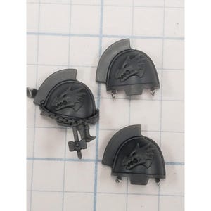 May include: Three gray plastic shoulder pads with a dragon emblem. One pad has a chain and a small hammer attached. The pads are designed for miniature figures and are likely for a tabletop game.