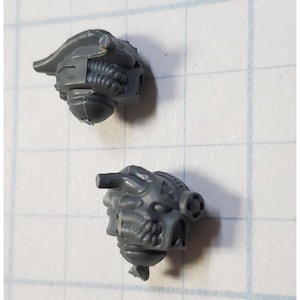 May include: Two gray plastic miniature heads with detailed sculpting. The heads have a futuristic design with pipes and wires.