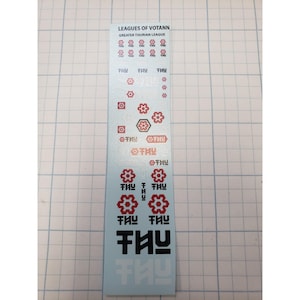 May include: A light blue sticker sheet with the text "LEAGUES OF VOTANN GREATER THURIAN LEAGUE" in black. The sheet features red and white graphic designs, including flower-like shapes and stylized text. The background is a grid.