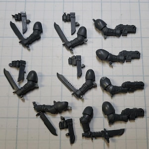 May include: A collection of unpainted, gray plastic miniature arms, weapons, and accessories. The assortment includes swords, bolters, and pouches, likely for a tabletop war game. The pieces are arranged on a grid-lined surface.